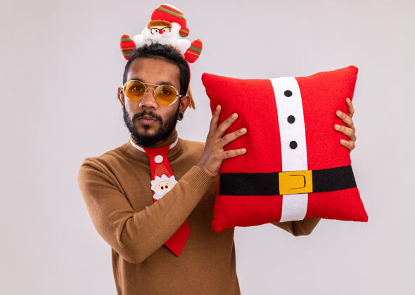 轮辋一个穿着棕色毛衣 头戴圣诞老人戒指 打着有趣的红色领带的非洲裔美国人拿着圣诞枕头 站在白色背景下看着相机 微笑着自信地站着枕头圣诞节自信