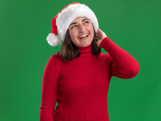 帽子穿着红毛衣 戴着圣诞帽的年轻女孩面带微笑 站在绿色的墙边 快乐而快乐微笑年轻毛衣