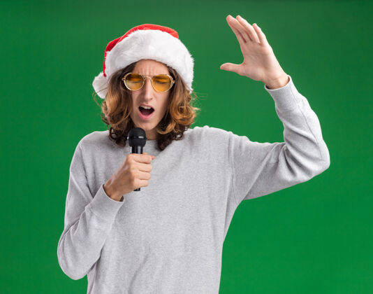 圣诞节一个戴着圣诞老人帽和黄色眼镜的情绪化的年轻人举起手臂站在绿色背景上对着麦克风大声喊叫举起麦克风手臂