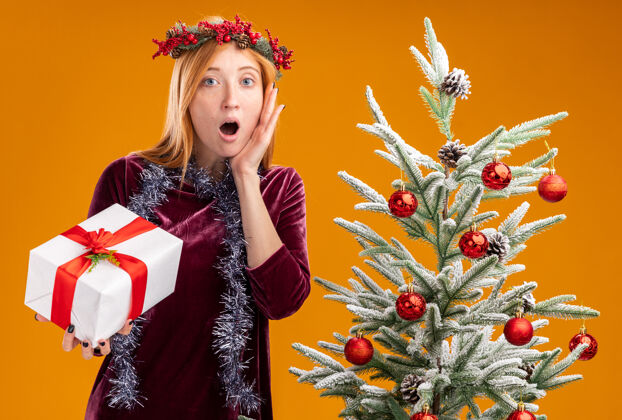 手站在圣诞树旁的年轻漂亮女孩 身穿红色连衣裙 脖子上戴着花环 手里拿着礼盒 手放在脸颊上 孤立在橙色背景上抱着年轻衣服