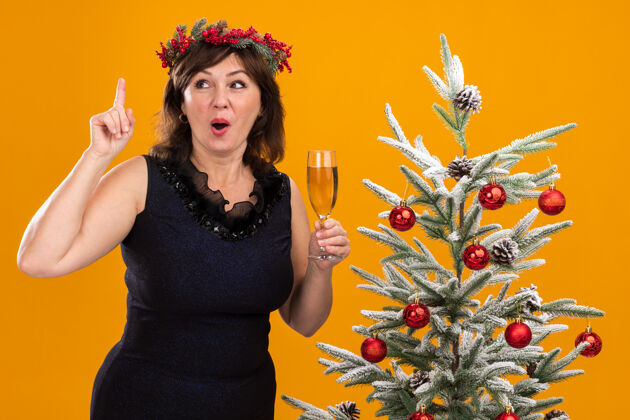 靠近令人印象深刻的中年妇女头戴圣诞花环 脖子上戴着金箔花环 站在装饰过的圣诞树旁 手里拿着一杯香槟向上围着站着
