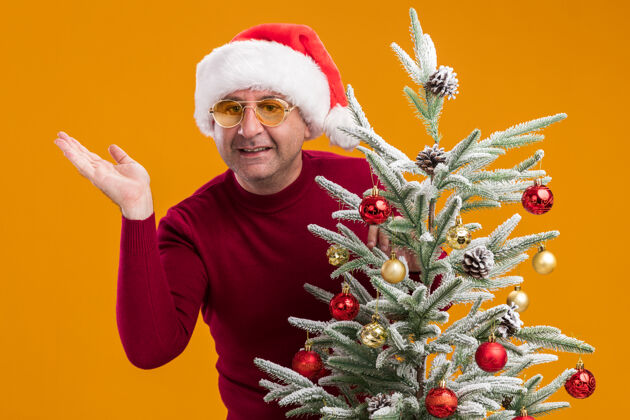深色中年男子戴着圣诞老人帽 穿着深红色高领毛衣 戴着黄色眼镜 装饰着圣诞树 站在橙色的背景下 看着摄像机兴高采烈地微笑着帽子微笑橙色