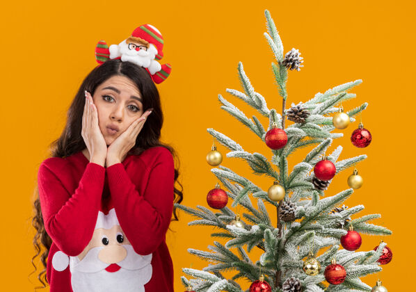 橙色困惑的年轻漂亮女孩戴着圣诞老人的头带和毛衣站在装饰过的圣诞树旁手放在脸上看着摄像机噘着嘴唇隔离在橙色背景下年轻圣诞树漂亮
