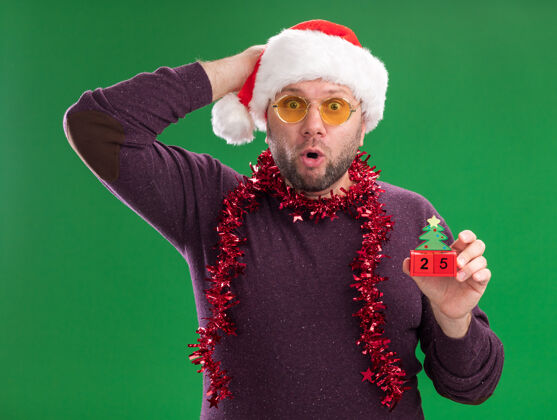头一个戴着圣诞帽 脖子上戴着金属丝花环 戴着眼镜 头上拿着圣诞树玩具 头上戴着枣子的中年男人看着隔离在绿色背景上的相机眼镜中年关心