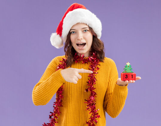 举行惊讶的年轻斯拉夫女孩戴着圣诞帽 脖子上戴着花环 拿着圣诞树的装饰物 在紫色的背景上孤立着复制空间脖子指着圣诞老人