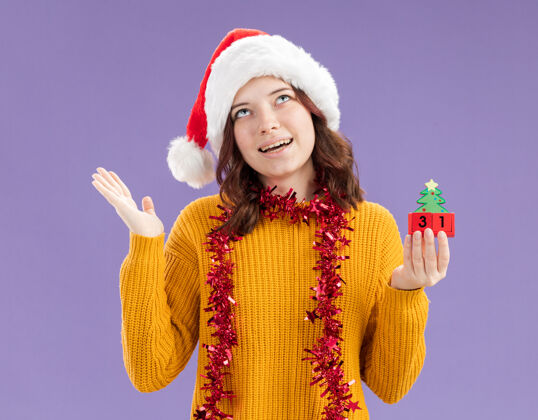 花环年轻的斯拉夫女孩戴着圣诞帽 脖子上戴着花环 手拿圣诞树饰物 抬头仰望着紫色背景上与世隔绝的复制空间女孩圣诞周围