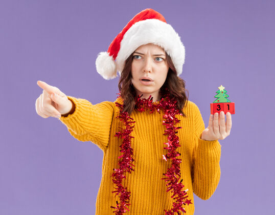 圣诞老人戴着圣诞帽 脖子上戴着花环的年轻斯拉夫女孩拿着圣诞树饰物 看着并指着在紫色背景上有复制空间的隔离侧圣诞树装饰品举行