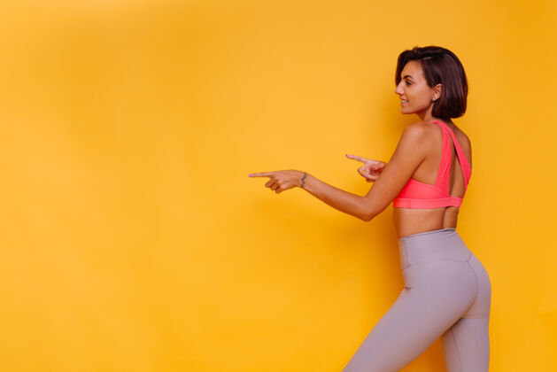 时尚模特年轻健壮的女人穿着运动服 时髦的上衣和紧身裤 对着黄色的墙壁摆姿势动机积极强壮