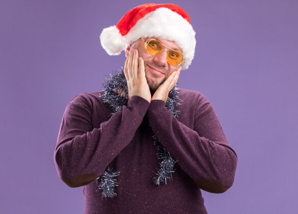 圣诞老人高兴的中年男子戴着圣诞帽 脖子上戴着金属丝花环 戴着眼镜 手放在脸上 看着紫色背景上孤立的摄像机手帽子脖子