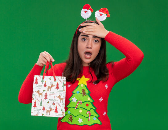 头带带着圣诞老人头带的焦急的年轻白人女孩把手放在额头上 拿着一个纸礼品袋 隔离在绿色背景上 留有复印空间包年轻圣诞老人