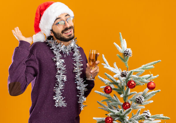 花环微笑的年轻帅哥站在圣诞树旁 戴着圣诞帽 脖子上戴着花环 手里拿着一杯橘黄色背景的香槟小伙子抱着香槟