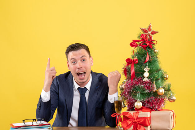 礼物正面图惊奇的男人手指着坐在圣诞树旁的桌子和黄色墙上的礼物指着惊奇的男人男人