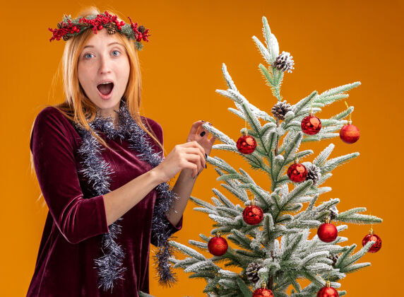 站着站在圣诞树旁的年轻漂亮的女孩穿着红色的裙子 脖子上戴着花环 橙色的背景下孤立着女孩圣诞节衣服