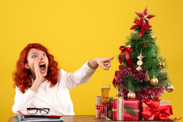 情感女工人坐在桌子后面 手里拿着圣诞礼物和黄色的圣诞树坐着工人礼物
