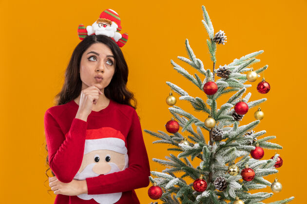 圣诞老人体贴的年轻漂亮女孩 戴着圣诞老人的头带和毛衣 站在装饰好的圣诞树旁 手放在下巴上 在橙色的背景下孤立地抬头看近橙色装饰