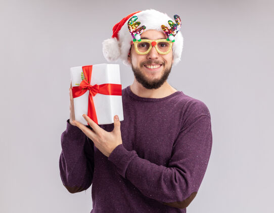 搞笑穿着紫色毛衣 戴着圣诞帽 戴着滑稽眼镜的快乐的年轻人拿着礼物 站在白色的背景下 兴高采烈地微笑着看着镜头年轻礼物抱着