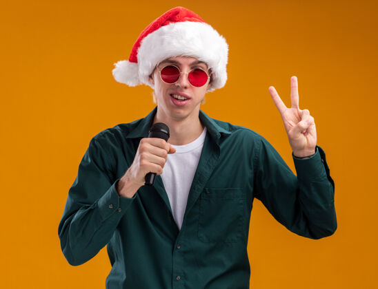 眼镜自信的年轻金发男子戴着圣诞帽和眼镜对着麦克风说话看着摄像机在橙色背景上做和平标志帽子自信年轻