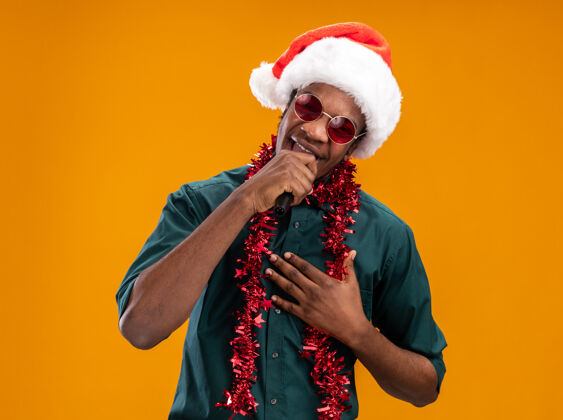 圣诞老人戴着圣诞帽 戴着花环 戴着眼镜的非裔美国人站在橙色背景下 对着麦克风快乐地唱歌麦克风眼镜站着
