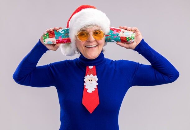微笑戴着太阳眼镜 戴着圣诞帽 系着圣诞领带 面带微笑的老妇人把纸杯举近耳朵 在白色背景上隔离开来 留有复印空间领带抱着亲密