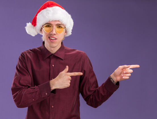 指着令人印象深刻的年轻金发男子戴着圣诞帽和眼镜看着摄像机指着一边孤立的紫色背景年轻印象深刻眼镜