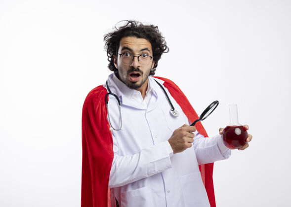 穿着一个戴着眼镜 身穿医生制服 披着红色斗篷 脖子上戴着听诊器的白人年轻人 手里拿着放大镜和玻璃瓶中的红色化学液体液体眼镜年轻