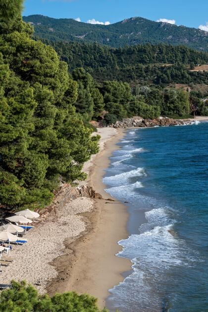平静希腊的爱琴海海岸 长满树木和灌木丛的岩石小山 有海浪的海滩和带日光浴床的雨伞蓝色度假村户外
