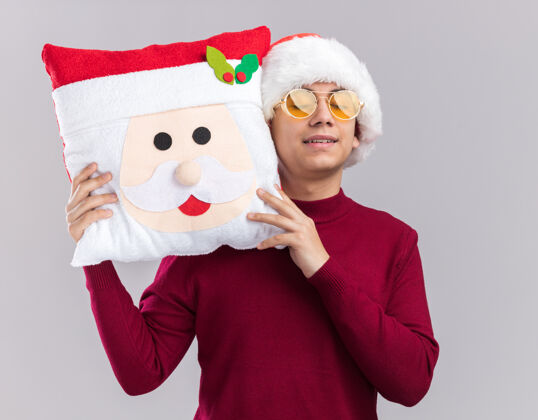 闭上喜欢闭着眼睛的年轻人戴着圣诞帽戴着眼镜抱着圣诞枕头隔离在白色背景上枕头圣诞节帽子
