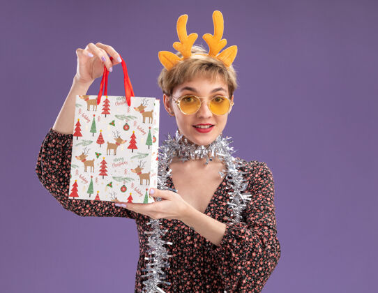 花环年轻漂亮的女孩戴着驯鹿鹿角头带 脖子上戴着金箔花环 戴着眼镜 手里拿着圣诞礼品袋 看着相机 背景是紫色 有复制空间金属丝年轻眼镜