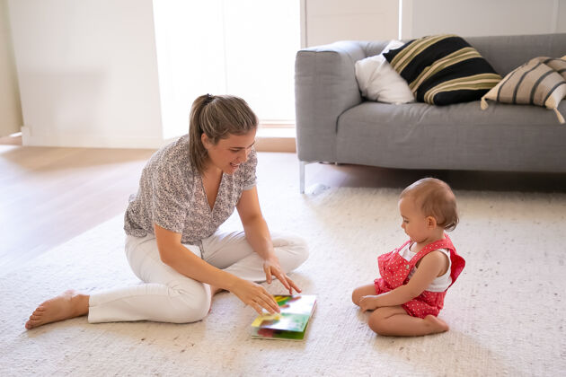 孩子漂亮的妈妈在给穿着红色短裙的可爱的小宝宝读书专心的孩子坐在客厅的地毯上学习阅读家庭 母亲和在家的概念生活年轻爱
