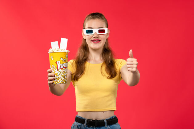 成人前视图年轻女性手持爆米花包 戴着d型太阳镜 站在浅红墙上的电影院里年轻女性电影女性