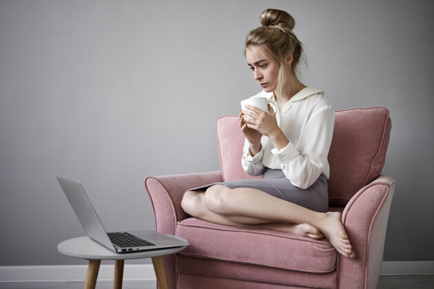 笔记本电脑照片中 穿着裙子和白衬衫的年轻金发白种女人光着脚 一边喝着大杯咖啡 一边在普通笔记本电脑上观看严肃的在线节目女性椅子坐着