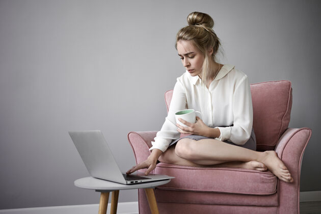 沙发漂亮的女性自由职业者 穿着白色上衣 在便携式电脑上远程工作 光着脚坐在扶手椅上 喝着热茶或咖啡 一只手放在键盘上 神情专注室内窗户休闲