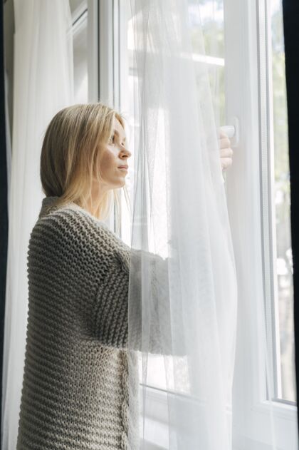 流行病大流行期间悲伤的女人在家的侧视图透过窗户看隔离垂直隔离