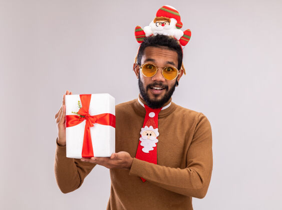 圣诞老人快乐的非洲裔美国人 身穿棕色毛衣 头戴圣诞老人戒指 打着有趣的红色领带 拿着礼物 站在白色背景下 面带微笑地看着相机毛衣站着轮辋