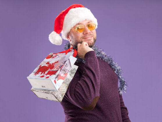脖子高兴的中年男子戴着圣诞帽 脖子上戴着金箔花环 戴着眼镜站在侧视图上 肩上扛着圣诞礼品袋 看着紫色背景上孤立的摄像机圣诞节周围圣诞老人