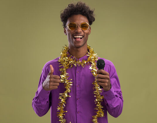 快乐快乐的非洲裔美国年轻人戴着眼镜 脖子上戴着金箔花环 手持麦克风 看着相机 在橄榄绿的背景上孤立地竖起大拇指眼镜金属片年轻