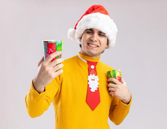圣诞老人快乐的年轻人穿着黄色高领毛衣 戴着圣诞老人帽 打着滑稽的领带 手里拿着五颜六色的纸杯 站在白色的背景上微笑着看着纸杯领带搞笑年轻