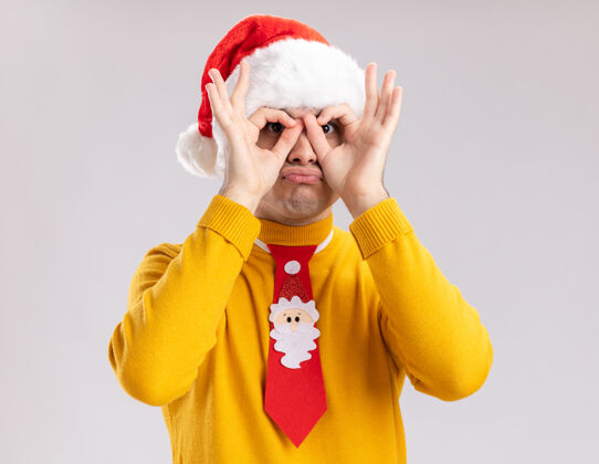 制作滑稽快乐的年轻人穿着黄色高领毛衣 戴着圣诞老人帽 戴着滑稽的领带 站在白色的背景上 用手指做着双目手势搞笑手指圣诞老人