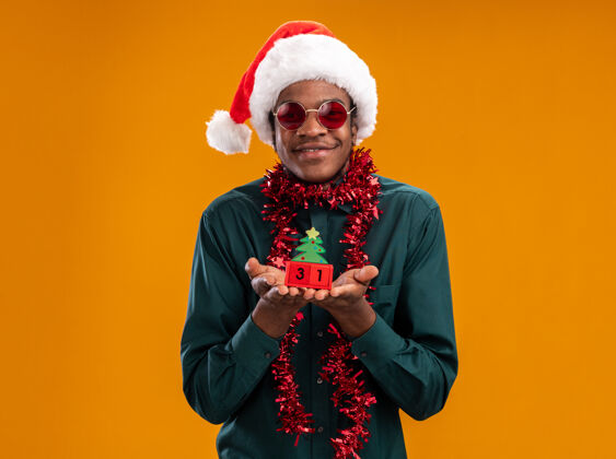 花环快乐的非洲裔美国人戴着圣诞帽 戴着花环 戴着太阳镜 抱着新年约会的玩具立方体 站在橙色背景下 看着相机 开心地微笑着立方体高兴美国