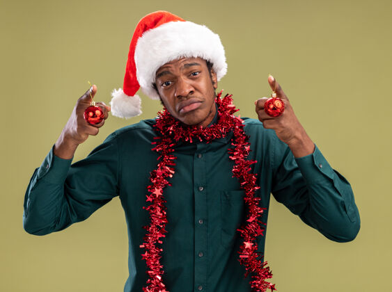 举行一个戴着圣诞帽 戴着花环 拿着圣诞球的非洲裔美国人站在绿色背景下 愁眉苦脸地撅着嘴唇看着摄像机花环圣诞老人站立