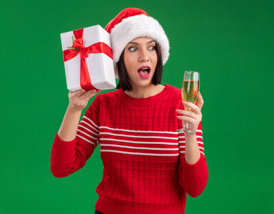 年轻好奇的年轻女孩戴着圣诞帽拿着礼包靠近头部和一杯香槟看着绿色背景上孤立的一面帽子持有女孩