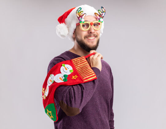 帽子快乐快乐的年轻人穿着紫色毛衣 戴着圣诞帽 戴着滑稽的眼镜 手里拿着圣诞长袜 站在白色的背景下微笑着看着镜头圣诞节毛衣袜子