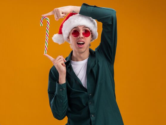 帽子令人印象深刻的年轻金发男子戴着圣诞帽和眼镜手持圣诞甜手杖垂直看着相机孤立的橙色背景与复制空间金发垂直圣诞老人