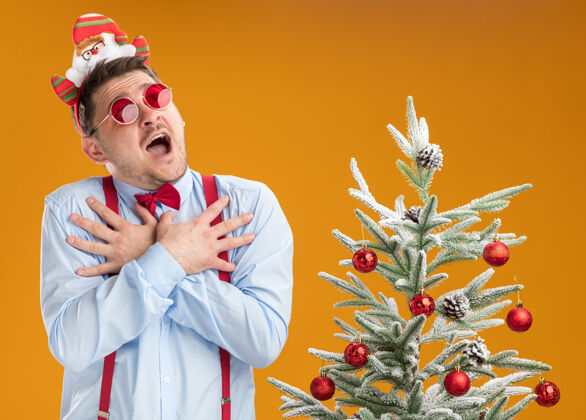 胸部戴着吊带领结的年轻人站在圣诞树旁 戴着圣诞老人和红眼镜 在橙色的背景下 手放在胸前 惊讶而担忧地抬头望着吊带手圣诞老人
