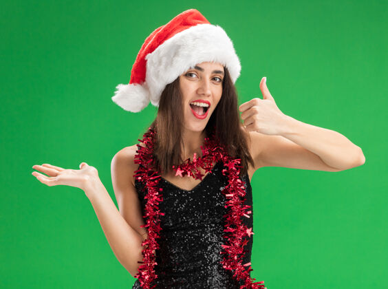 拇指快乐的年轻漂亮女孩戴着圣诞帽 脖子上戴着花环 大拇指向上张开 手孤立在绿色背景上女孩花环圣诞节