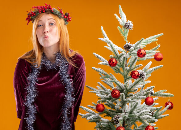 圣诞站在圣诞树旁的年轻漂亮女孩 身穿红色连衣裙 脖子上戴着花环 在橙色背景上显示出孤立的亲吻手势女孩花环年轻