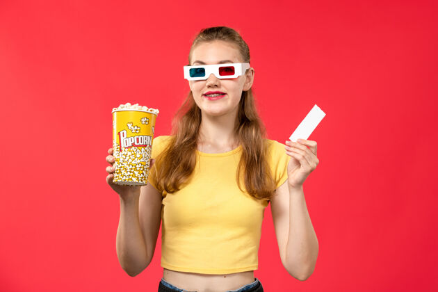 微笑前视图年轻女性手持爆米花包和票在d太阳镜在淡红墙影院电影年轻女性门票护目镜