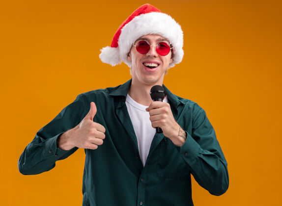 圣诞老人快乐的年轻金发男子戴着圣诞帽 戴着眼镜 拿着麦克风 看着相机 在橙色背景上孤立地竖起大拇指眼镜金发拿着