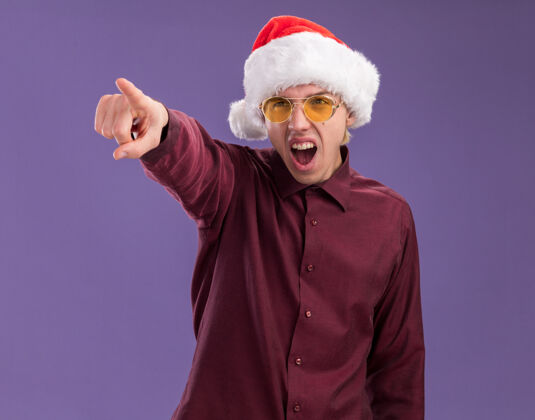 眼镜年轻的金发男子戴着圣诞帽和眼镜看着并指着紫色背景上孤立的一面年轻圣诞老人金发