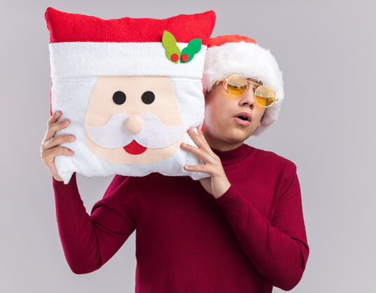 帽子惊讶的年轻人戴着圣诞帽戴着眼镜拿着圣诞枕头隔离在白色背景上圣诞节小伙子抱着